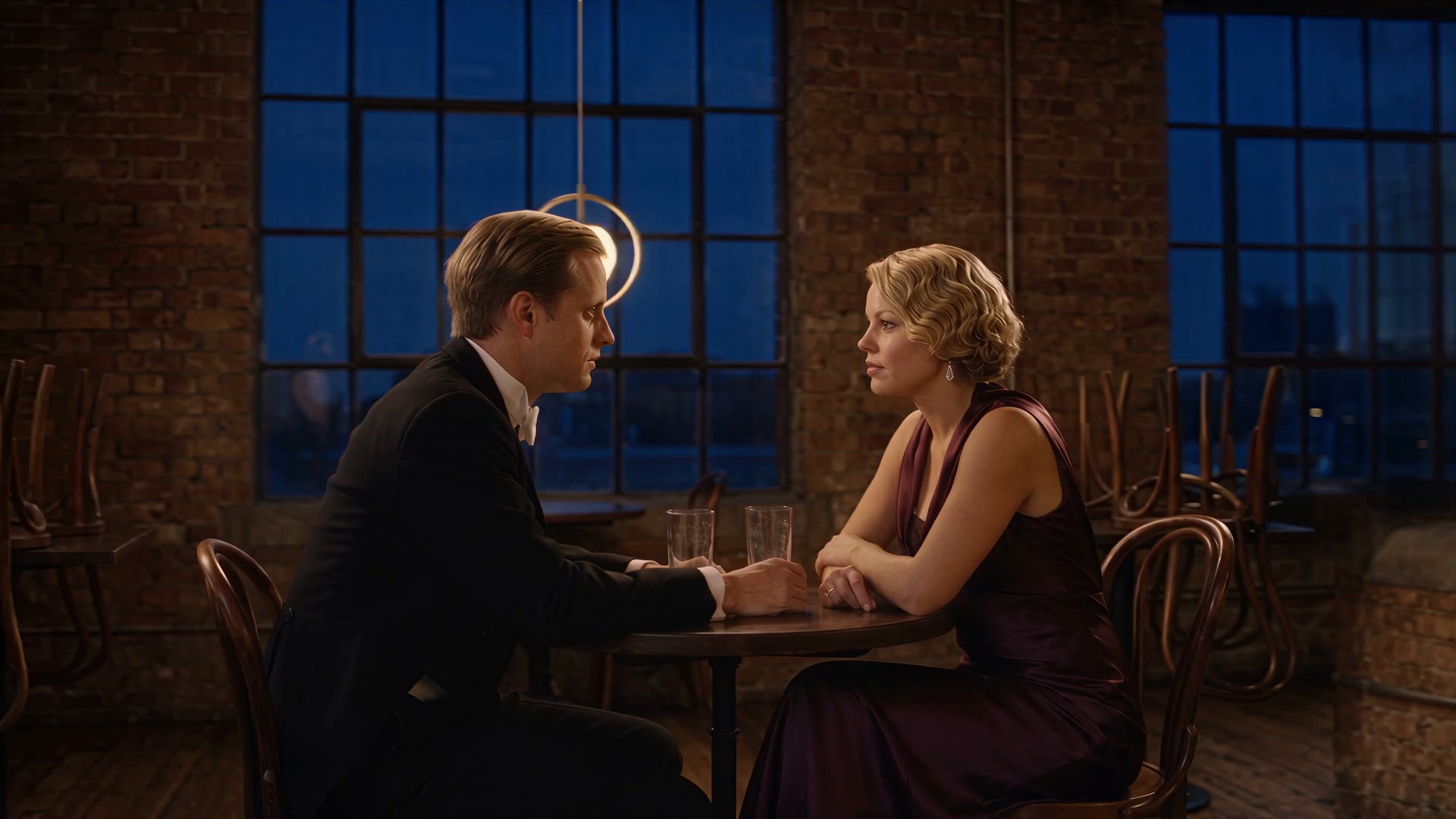 Two people at a small table by a tall industrial window at dusk, hands almost touching.